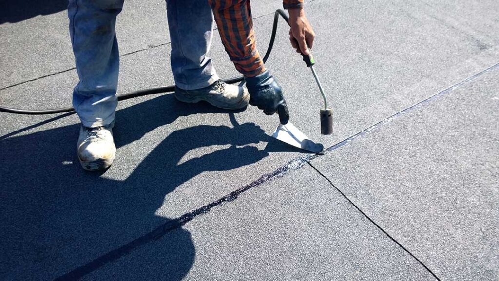 Roofers inspect new roofing above a home while dry attic insulation and roof framing are visible below.