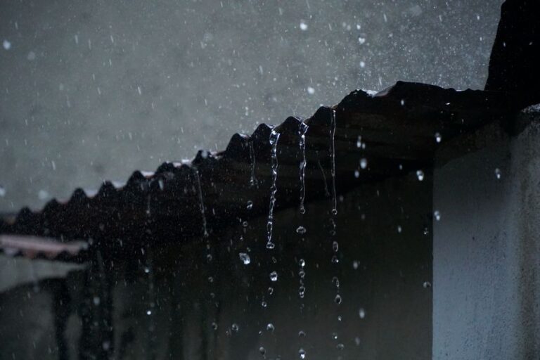 A homeowner examines a ceiling water stain beneath a roof leak while rain falls outside.