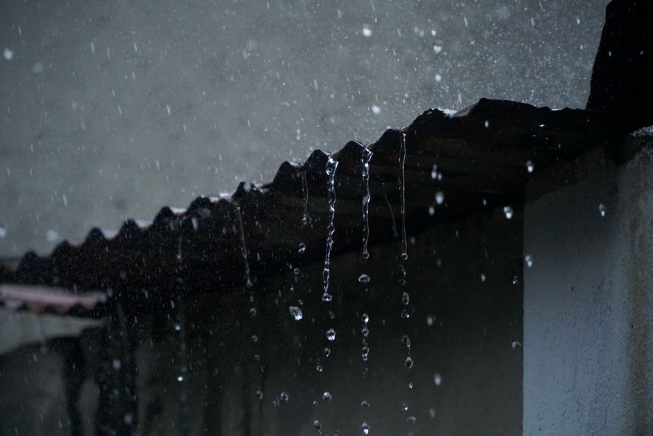 A homeowner examines a ceiling water stain beneath a roof leak while rain falls outside.